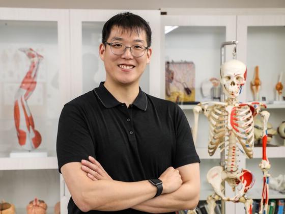 Photo of an Asian male in a black shirt standing in an anatomy lab with a model skeleton to his right.
