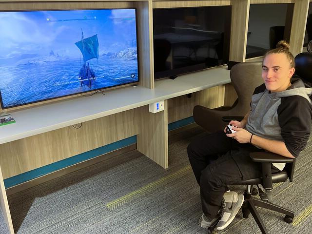 一名男学生拿着电子游戏控制器，对着大屏幕上播放电子游戏的摄像头微笑.